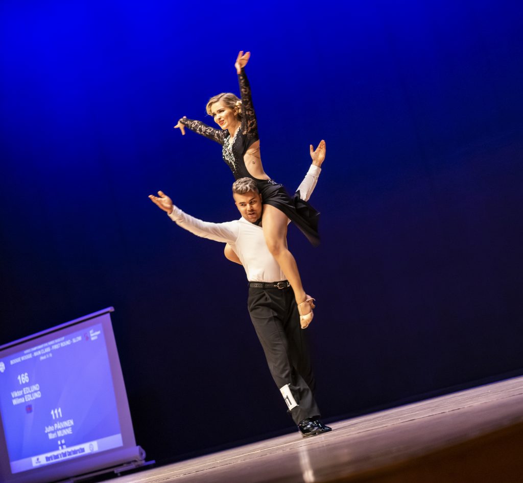 Juho Päivinen ja Mari Munne tanssimassa. Mari istuu Juhon olkapäällä, molemmilla kädet kohotettuna yläviistoon.