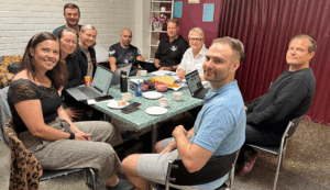 Miehiä ja naisia istuu pöydän ympärillä ja katsoo kameraan.