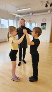 Hymyilevä nainen opastaa paritanssia tanssivaa lapsiparia.