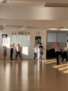 Lapsia seisomassa tanssisalin seinustalla, naisopettaja opettaa.