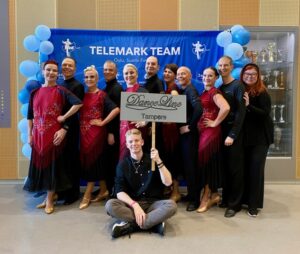 TAustalla ryhmä tanssijoita, edessä mies istuu seuran nimikyltin kanssa.